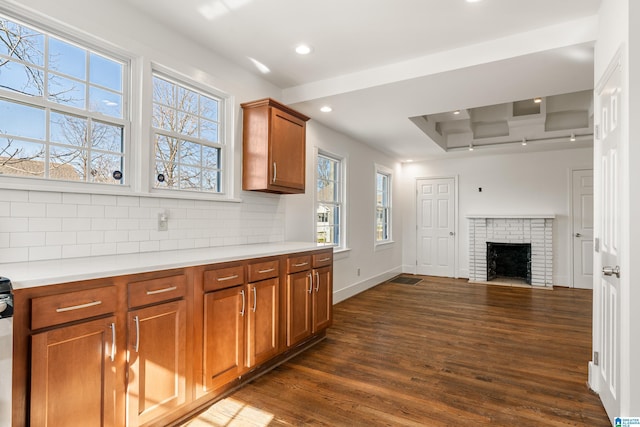 kitchen with light countertops, backsplash, brown cabinetry, dark wood-type flooring, and a brick fireplace