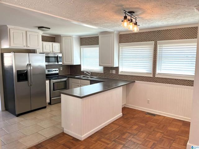 kitchen featuring dark countertops, a wainscoted wall, appliances with stainless steel finishes, a peninsula, and a sink