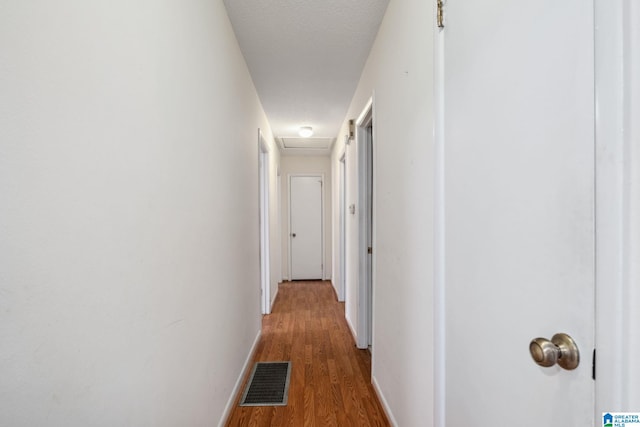 hallway featuring attic access, visible vents, baseboards, wood finished floors, and a textured ceiling