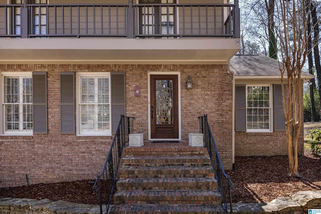 view of exterior entry with brick siding, a balcony, and roof with shingles