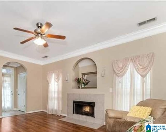 living area with ceiling fan, wood finished floors, visible vents, a tiled fireplace, and crown molding