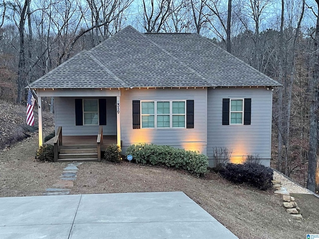 bungalow with covered porch, a wooded view, and roof with shingles