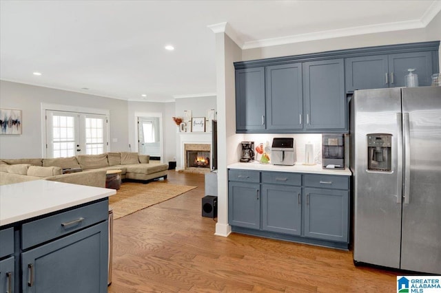 kitchen with a warm lit fireplace, stainless steel fridge with ice dispenser, ornamental molding, light countertops, and light wood-style floors