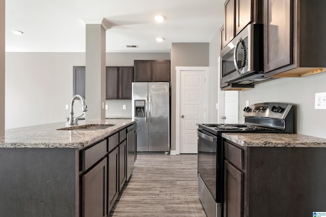 kitchen with light stone counters, stainless steel appliances, a sink, dark brown cabinetry, and light wood-type flooring