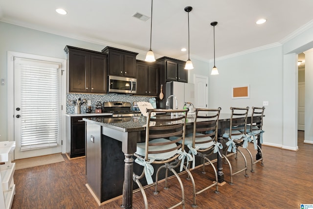 kitchen with a kitchen island with sink, ornamental molding, dark wood-style floors, stainless steel appliances, and decorative backsplash