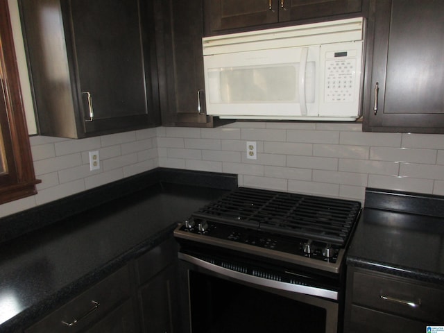 kitchen with white microwave, dark brown cabinetry, backsplash, dark countertops, and stainless steel range with gas stovetop