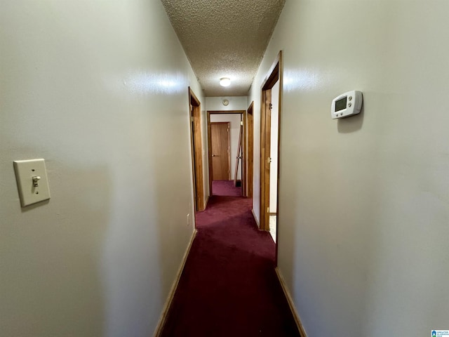 hall featuring a textured ceiling, dark colored carpet, and baseboards