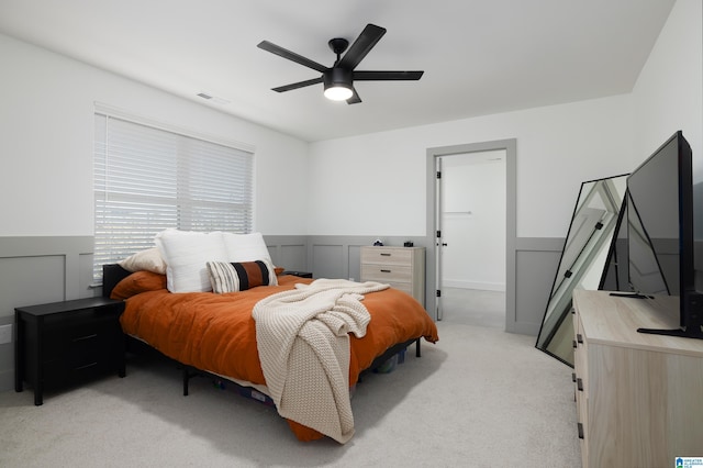 bedroom with light colored carpet, a decorative wall, visible vents, a ceiling fan, and wainscoting