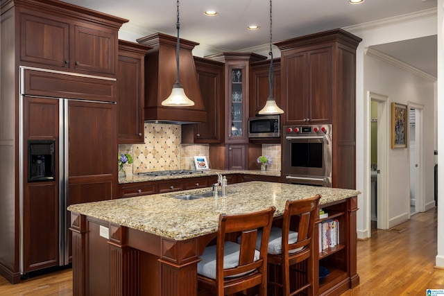 kitchen with built in appliances, light wood-style floors, hanging light fixtures, a center island with sink, and custom range hood