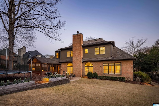 back of property at dusk featuring a trampoline, brick siding, a yard, and a chimney