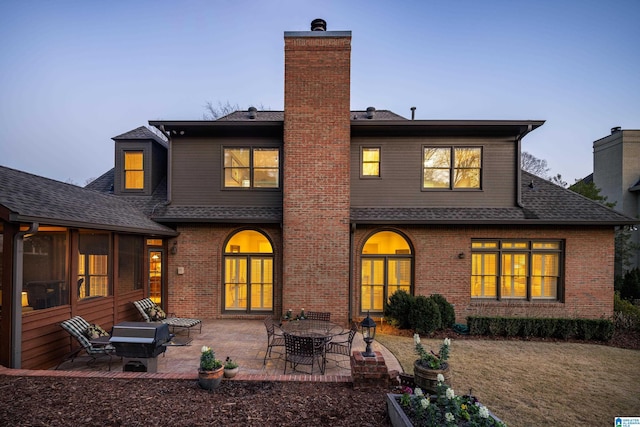 back of property at dusk with a patio area, a shingled roof, a chimney, and brick siding