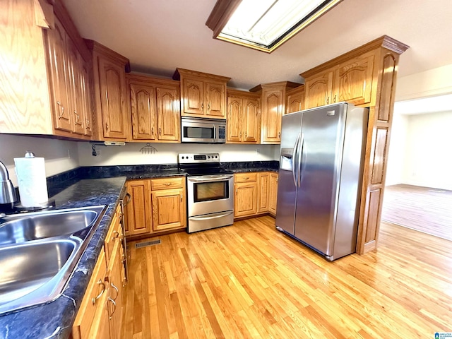 kitchen featuring light wood-style flooring, a sink, appliances with stainless steel finishes, brown cabinetry, and dark countertops