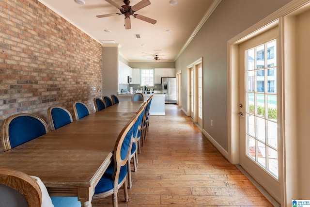 unfurnished dining area featuring ornamental molding, a ceiling fan, brick wall, light wood-type flooring, and baseboards