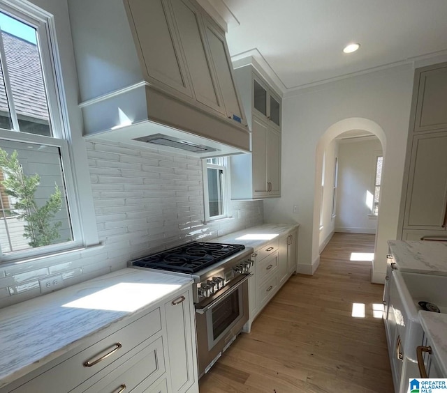 kitchen featuring arched walkways, stainless steel range, custom range hood, backsplash, and light wood-style flooring