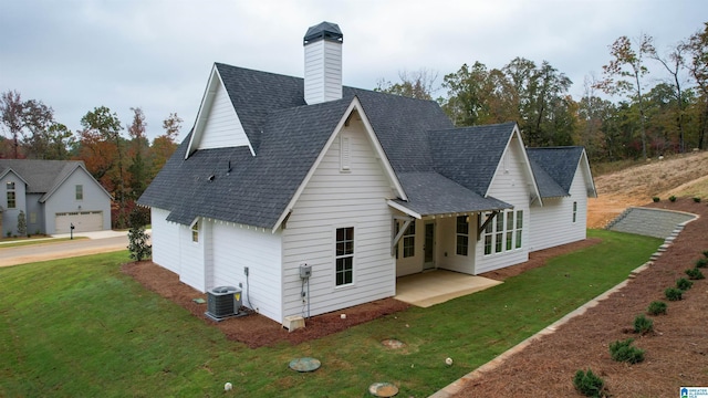 rear view of property with central AC unit, a shingled roof, a yard, a chimney, and a patio area