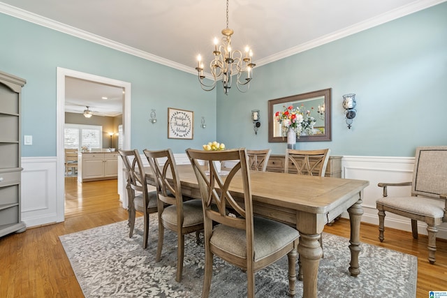 dining space featuring a wainscoted wall, ceiling fan with notable chandelier, light wood-type flooring, and crown molding