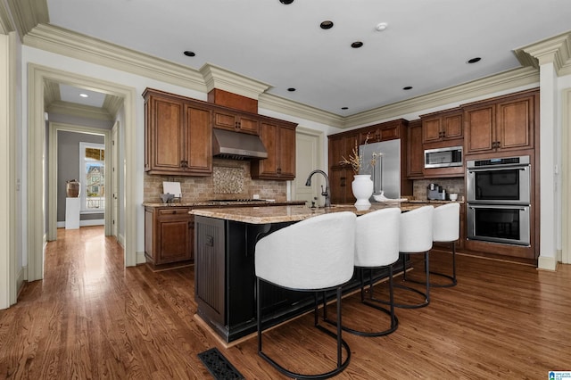 kitchen with under cabinet range hood, dark wood-style flooring, appliances with stainless steel finishes, tasteful backsplash, and a center island with sink