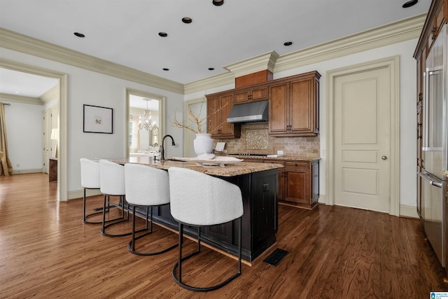 kitchen featuring dark wood-style floors, an island with sink, backsplash, and under cabinet range hood