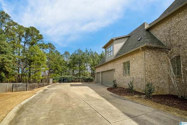 view of side of property with a garage, a shingled roof, brick siding, fence, and driveway