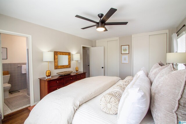 bedroom with a wainscoted wall, ensuite bath, dark wood-style flooring, multiple closets, and tile walls