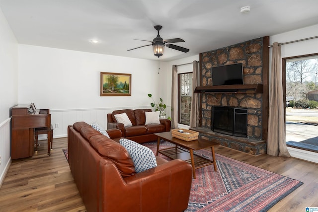 living area featuring a stone fireplace, wainscoting, wood finished floors, and a ceiling fan