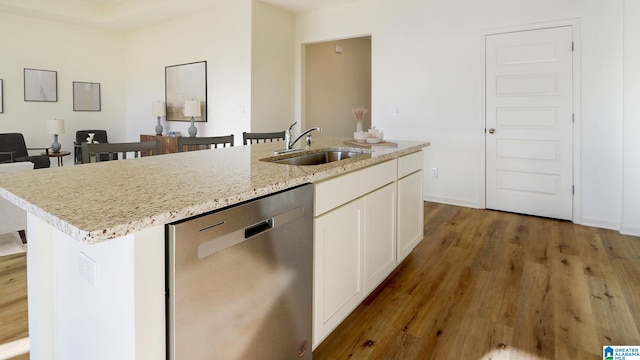 kitchen with wood finished floors, stainless steel dishwasher, a kitchen island with sink, a sink, and light stone countertops