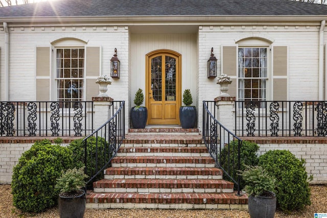 doorway to property featuring covered porch, brick siding, and roof with shingles