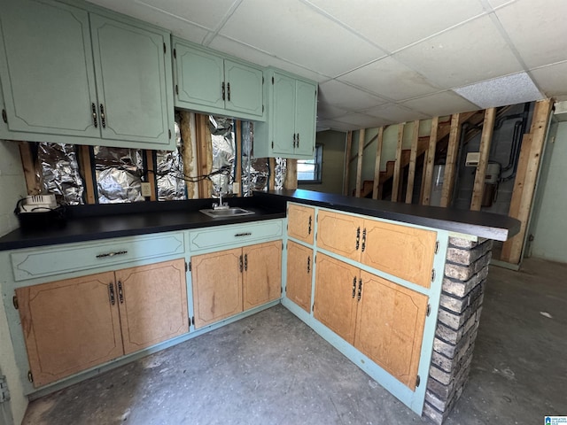 kitchen with dark countertops, a sink, concrete flooring, a drop ceiling, and a peninsula