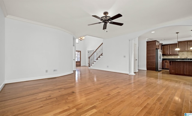 unfurnished living room with baseboards, ornamental molding, stairs, light wood-style floors, and ceiling fan with notable chandelier