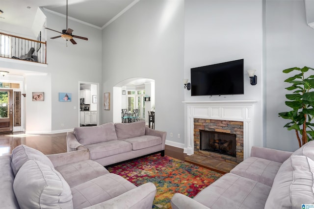 living area featuring arched walkways, a stone fireplace, wood finished floors, baseboards, and ornamental molding