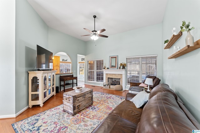 living room featuring wood finished floors, baseboards, a fireplace, arched walkways, and ceiling fan