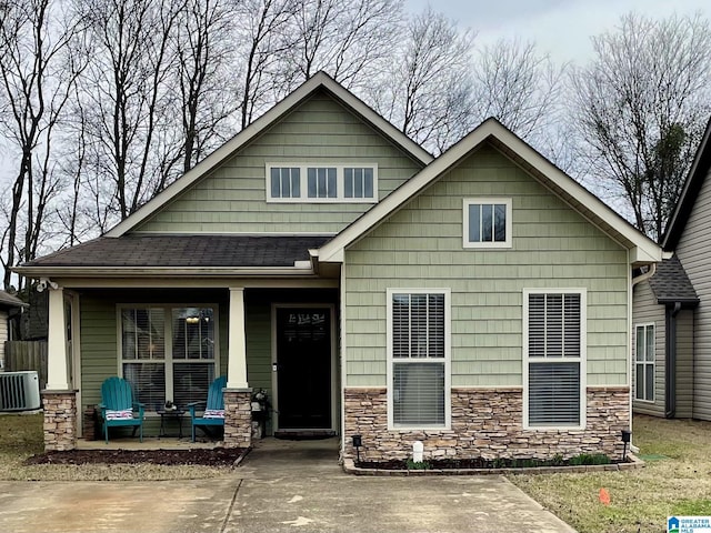 view of front of house featuring covered porch, stone siding, a shingled roof, and central air condition unit