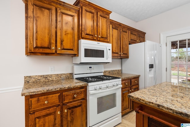 kitchen featuring a textured ceiling, white appliances, brown cabinets, and light stone countertops