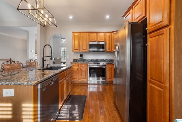 kitchen with stainless steel appliances, dark wood-style flooring, a sink, brown cabinetry, and dark stone countertops