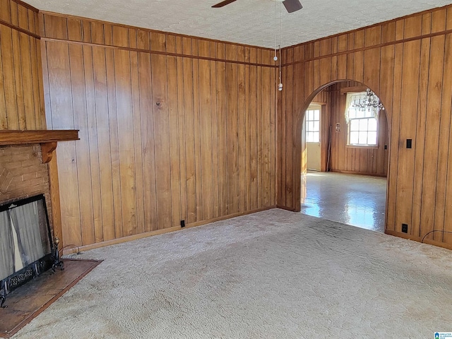 unfurnished living room with arched walkways, a ceiling fan, a brick fireplace, carpet flooring, and wooden walls