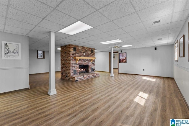 finished basement featuring a brick fireplace, visible vents, a drop ceiling, and wood finished floors