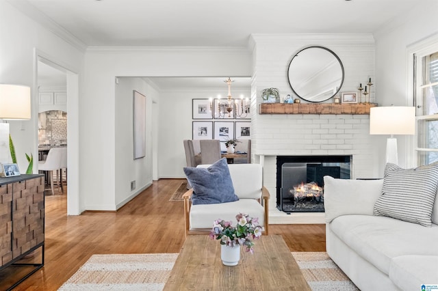 living area featuring wood finished floors, baseboards, crown molding, a brick fireplace, and a notable chandelier