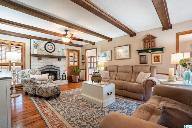 living room featuring ceiling fan, wood finished floors, a textured ceiling, a fireplace, and beam ceiling