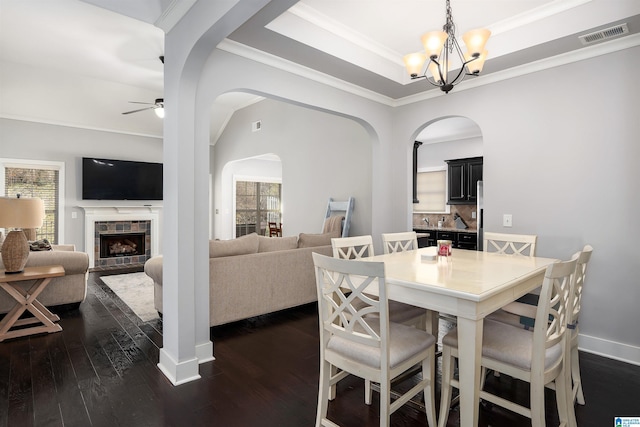 dining room with crown molding, dark wood-type flooring, a tiled fireplace, ceiling fan with notable chandelier, and a raised ceiling