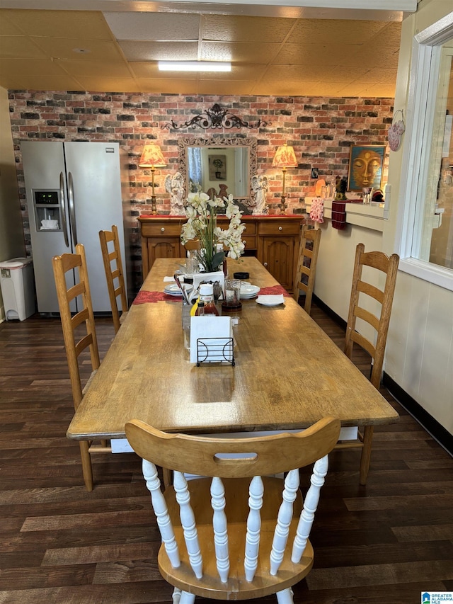 dining space with brick wall, baseboards, dark wood finished floors, and a drop ceiling