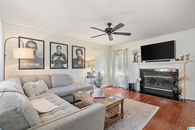 living room with ornamental molding, a ceiling fan, a tiled fireplace, wood finished floors, and baseboards