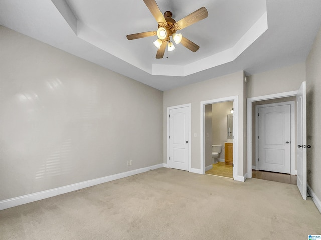 unfurnished bedroom featuring a ceiling fan, baseboards, ensuite bathroom, light carpet, and a raised ceiling