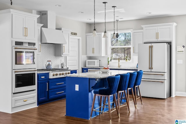 kitchen featuring a center island, blue cabinets, white cabinets, white appliances, and wall chimney exhaust hood