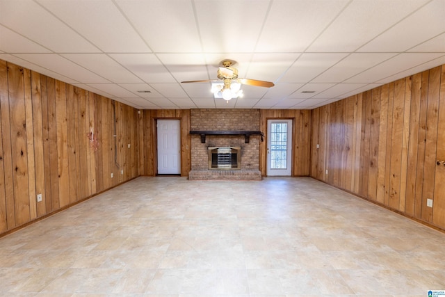 unfurnished living room featuring a brick fireplace, wood walls, ceiling fan, a drop ceiling, and baseboards