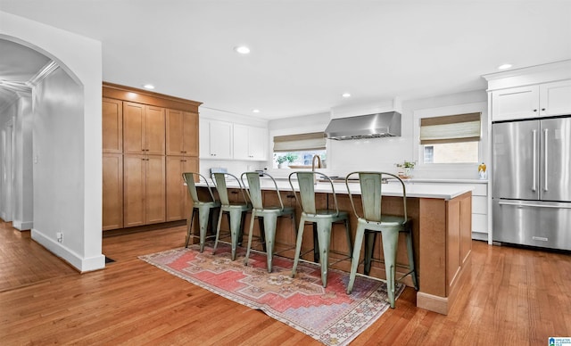 kitchen featuring a kitchen island with sink, light countertops, high end refrigerator, wall chimney range hood, and light wood-type flooring