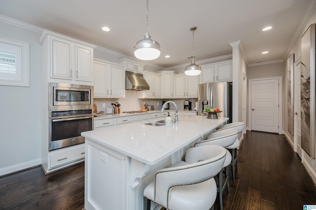 kitchen with appliances with stainless steel finishes, ornamental molding, a sink, under cabinet range hood, and a kitchen breakfast bar