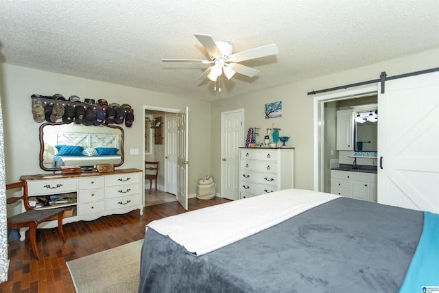 bedroom with a textured ceiling, ceiling fan, a barn door, dark wood-type flooring, and ensuite bath