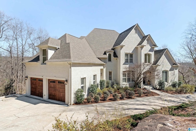 french provincial home with an attached garage, brick siding, driveway, and a shingled roof