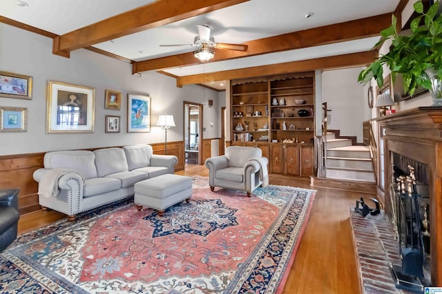 living room featuring beam ceiling, wood finished floors, stairway, a fireplace, and wainscoting