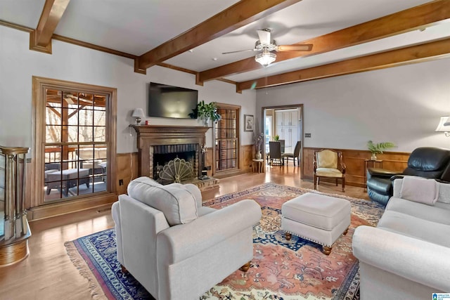 living room with a wainscoted wall, beam ceiling, a fireplace, wood finished floors, and a ceiling fan